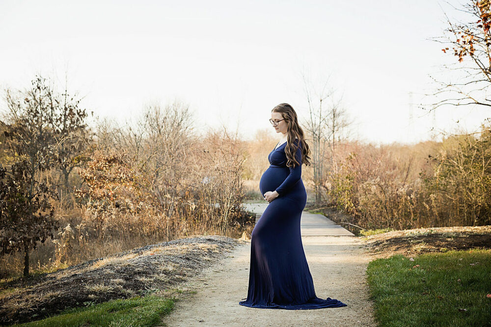 Woman doing pregnancy poses on park trail wearing long dress for her Navy maternity session taken South Jersey.