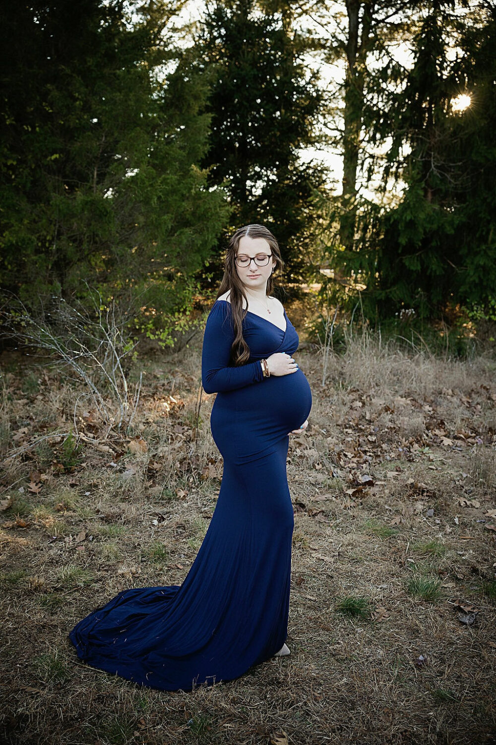 Woman holding her belly, wearing blue maternity dress, doing photography poses for her navy maternity session taken outdoors in Westhampton, New Jersey.