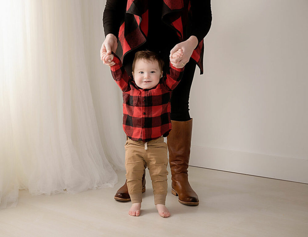 Toddler boy standing in front of his mother as she holds his arms up taken as a lifestyle portrait for his Wild ONe First Birthday sessions baby photos taken in Medford, New Jersey.