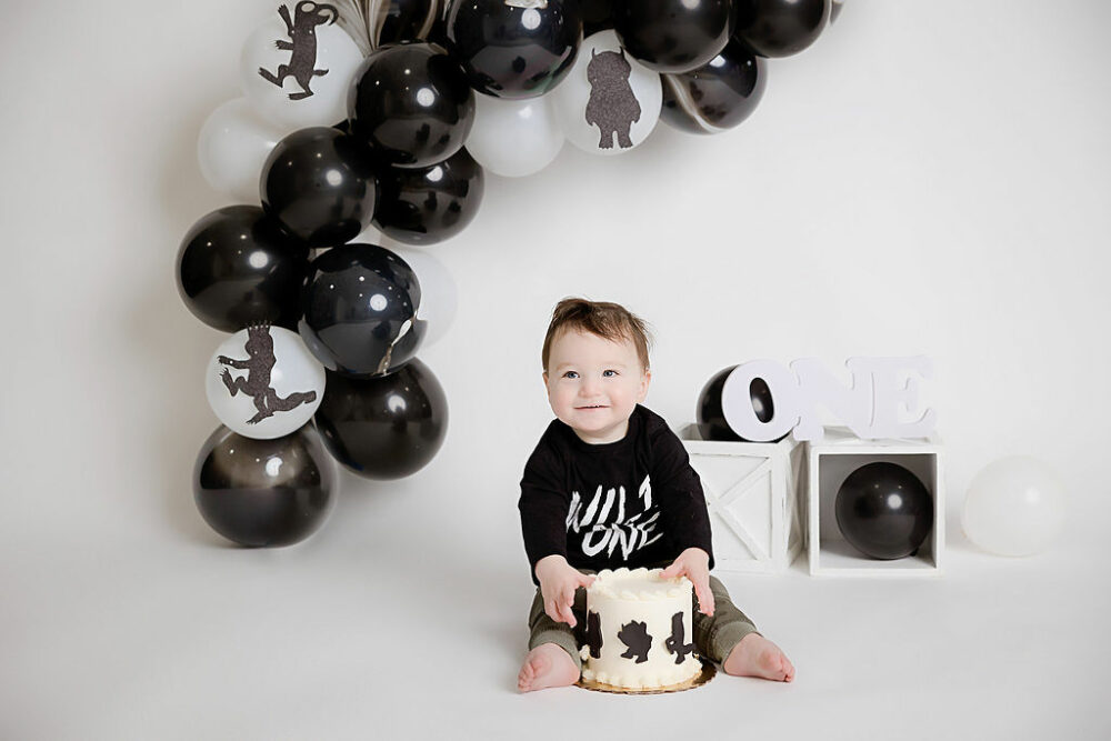 One year old boy sitting on floor behind a birthday cake with photography props and balloons as the background, smiling at the camera for his Wild one first birthday session taken in Southampton, New Jersey.