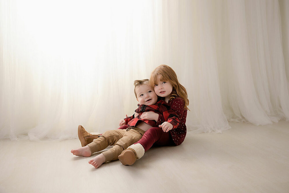 Toddler girl holding her one year old brother in her lap wearing matching clothes and sitting on the floor for his wWild one First Birthday session taken in studio in Monmouth, New Jersey.