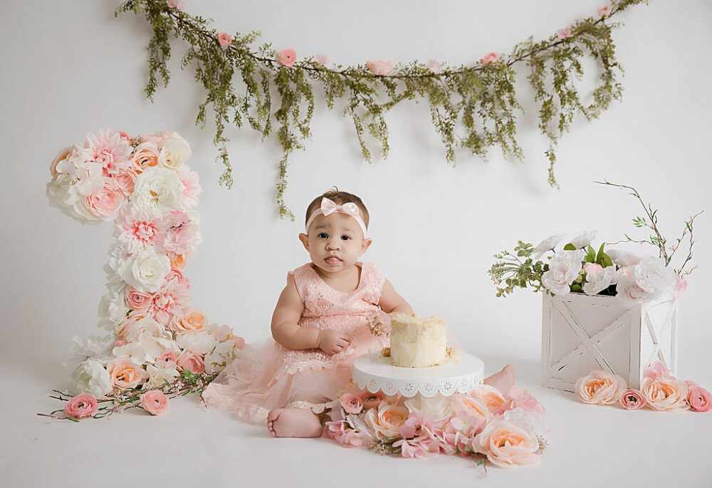 A one year old girl, looking at the camera as she sits on the floor, eating her first birthday cake with a theme of flowers and greenery, taken for her cake smash pictures in freehold, New Jersey.