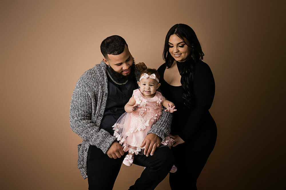 A family portrait of a father and mother, looking at their one year-old daughter for her studio First Birthday session taken in Marlton, New Jersey.