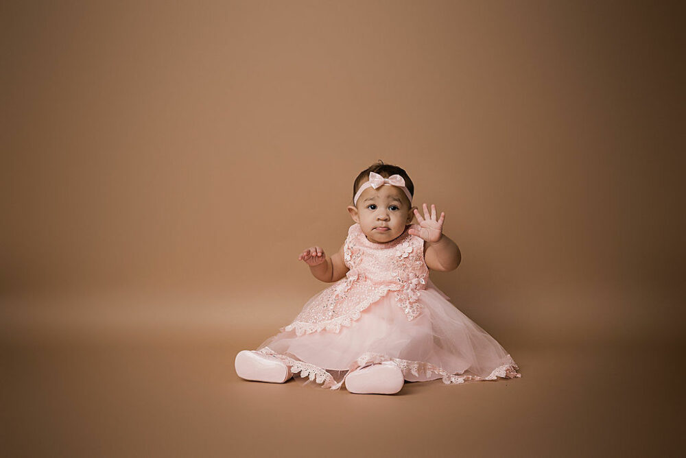 Toddler girl in pretty dress and matching headband is waving at the camera as she sits on the floor against backdrop for her moody first birthday session taken in Southampton, New Jersey.