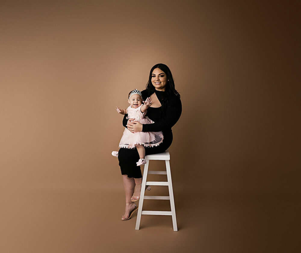 A woman sitting on ladder photography prop, while she hold her daughter on her lap, smiling at the camera for her in studio first birthday session, taken in Hamilton, New Jersey.