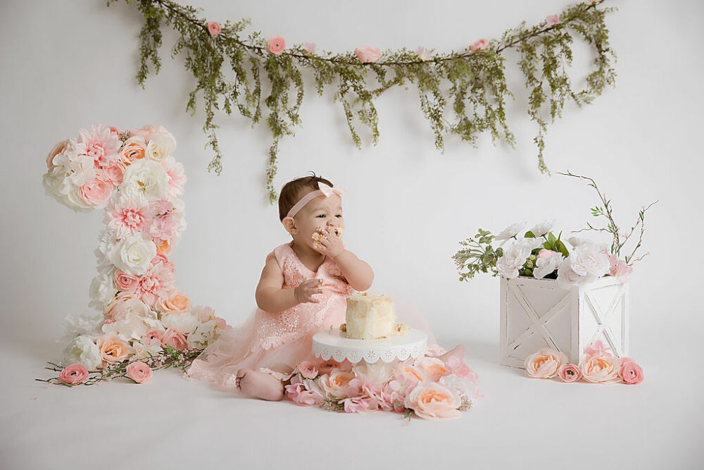 A toddler girl sitting on ground against a backdrop, eating cake, adorned with pink flowers for her cake, smash photos, taken in studio in Burlington, New Jersey.