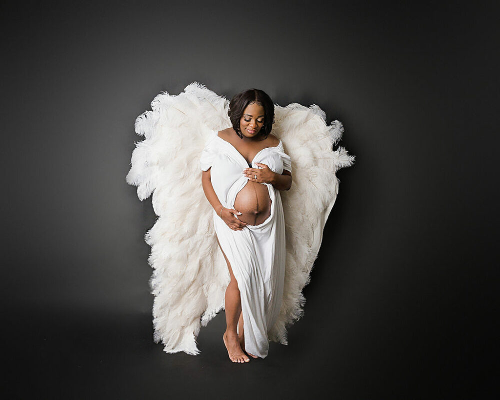 Best maternity photography of a woman wearing long gown with large angel wings for her and Maternity Photoshoot poses for her glamorous in-studio maternity session taken in Burlington, New Jersey.