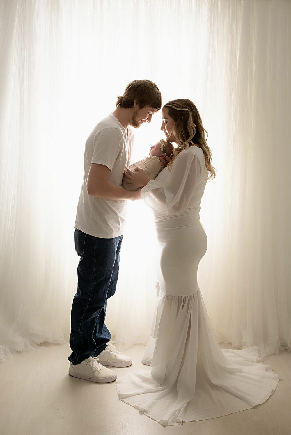 A man and a woman standing in facing each other holding their newborn in between them and almost touching foreheads for their floral newborn session taken in Mount Laurel, New Jersey.