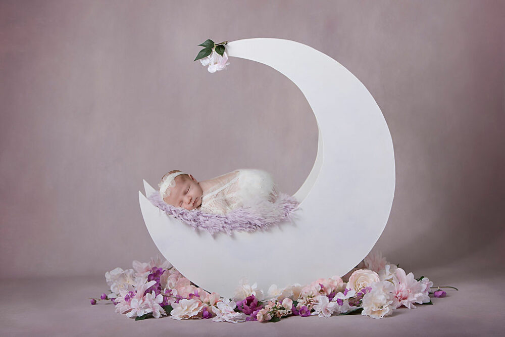 Newborn sleeping on textured blanket in moon shaped photography prop adorned with flowers for her floral newborn session in Southampton, New Jersey.