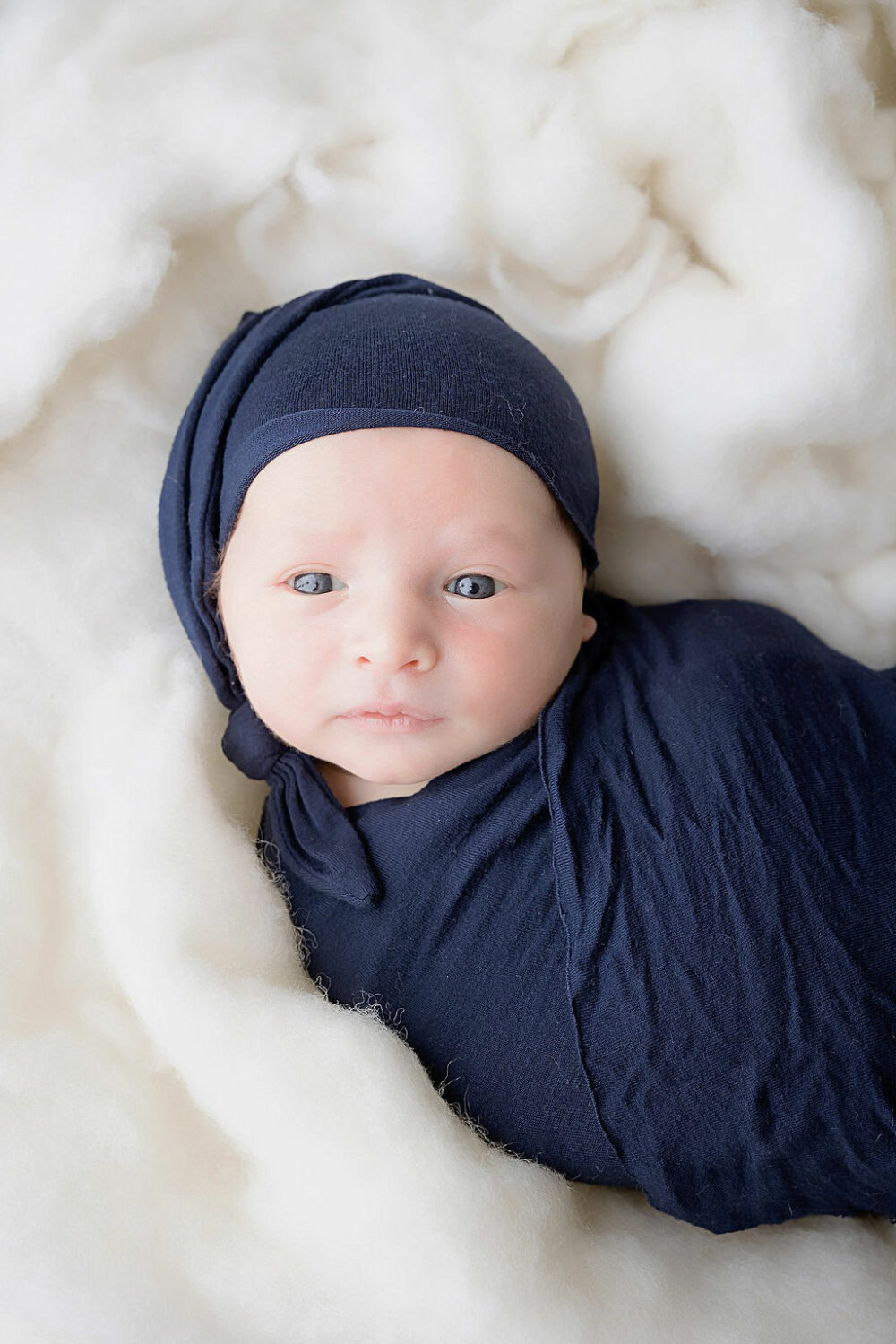 Close-up of infant boy laying on textured blanket swaddled, and wearing hat, looking at the camera for a navy newborn session, taken by professional photographer, in Cherry Hill, New Jersey.