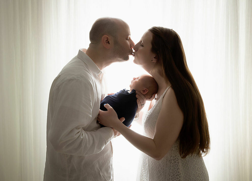 Man and woman facing each other and kissing while holding their newborn son between them against a light and bright background for their navy newborn session taken in Hamilton, New Jersey.