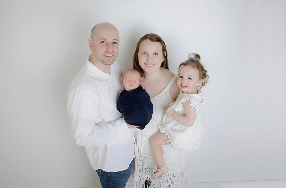 Family portrait of a man and a woman holding their newborn son in between them while mother hold their toddler girl for a Navy newborn session, taken in freehold, New Jersey.