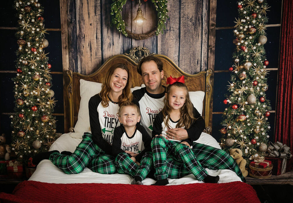 Holiday photo of a man and woman and her two toddler kids sitting on bed photography prop against a holiday backdrop for holiday mini sessions taken in Southampton, New Jersey.