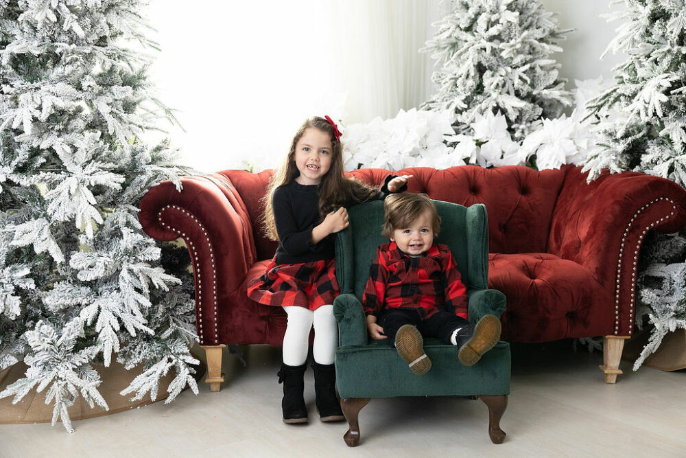 Toddler girl sitting on loveseat well her siblings it on a baby chair smiling a camera for their holiday portraits taken in Mount Holly, New Jersey.