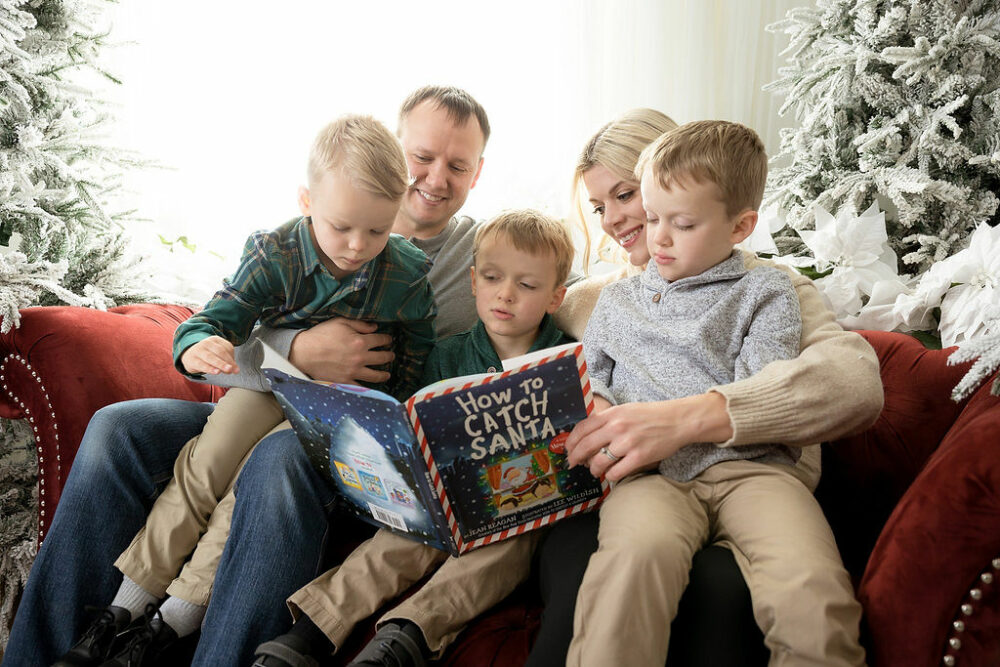 A man and woman sitting on loveseat reading to their three toddler boys posing for their family Christmas pictures taken in Mount Laurel, New Jersey.
