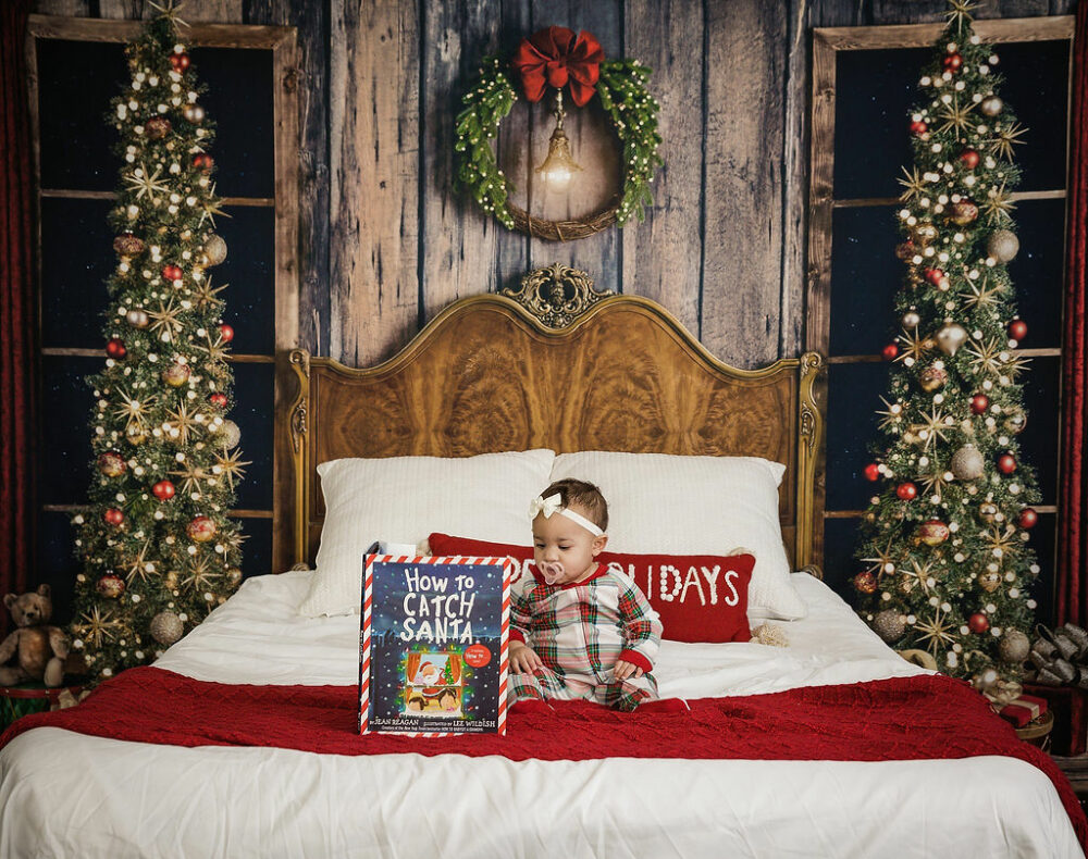 An infant girl wearing pajamas sitting on bed photography prop reading a book for her holiday mini session taken in studio in Warren, New Jersey.