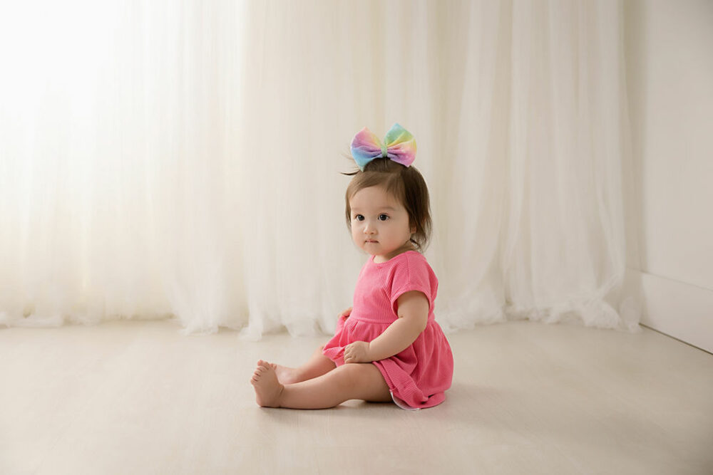 Toddler girl sitting on the floor wearing dress and a large headband taken during her baby milestones first birthday session taken in professional studio in Southampton, New Jersey.