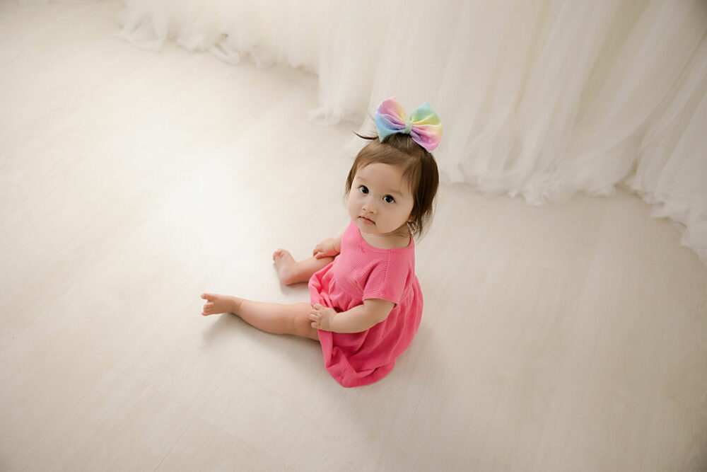 Toddler girl sitting and looking up at camera wearing dress and large headband on her head for her childhood milestones session taken in studio in Westampton, New Jersey.