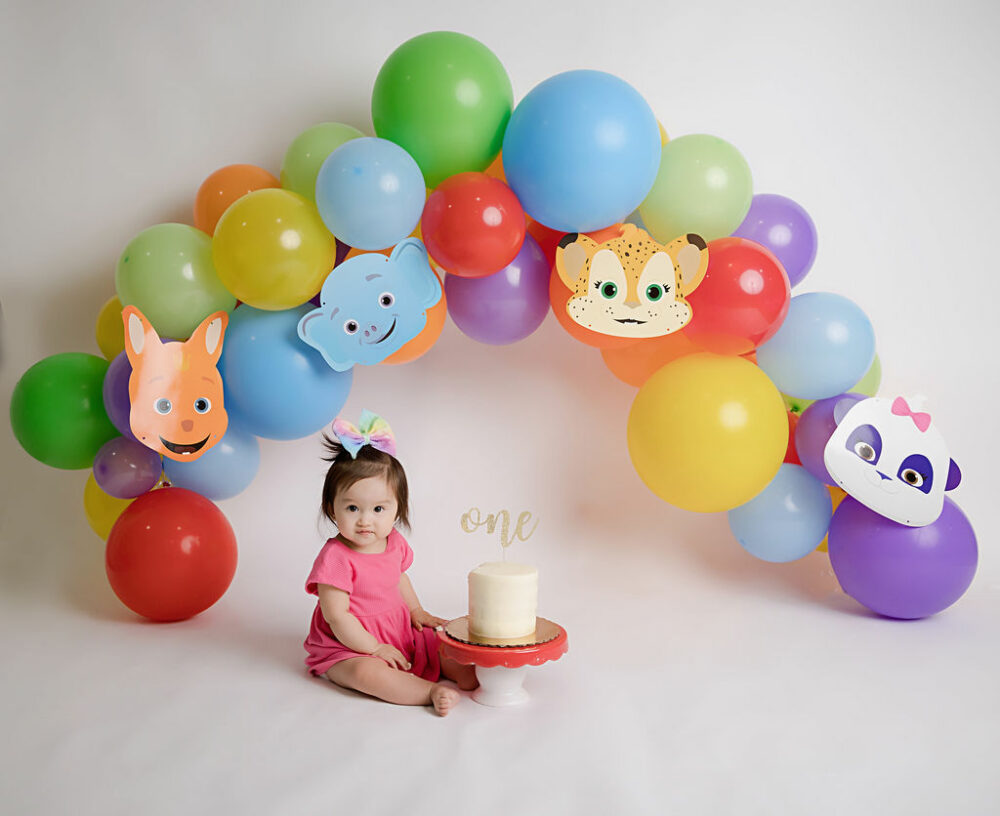 Toddler girl looking at camera and sitting next to a cake with balloons as the backdrop for her word party first birthday session taking an East Hampton, New Jersey.