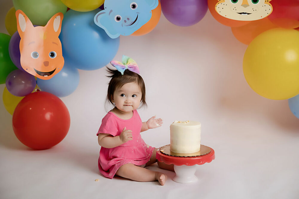 A close-up of a toddler girl sitting on the floor and eating her first birthday cake with word party characters in the backdrop taken in photography studio in Cherry Hill, New Jersey.