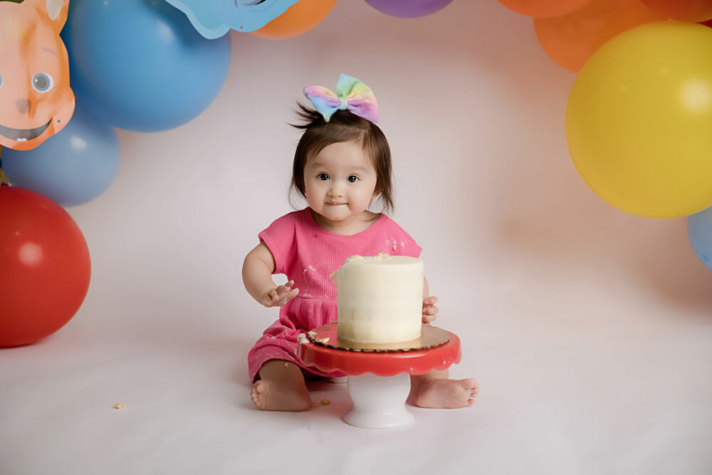 Toddler girl sitting behind photography prop with white cake on top, with her favorite word party characters and balloons as the backdrop for her baby milestones pictures taken in Mount Holly, New Jersey.