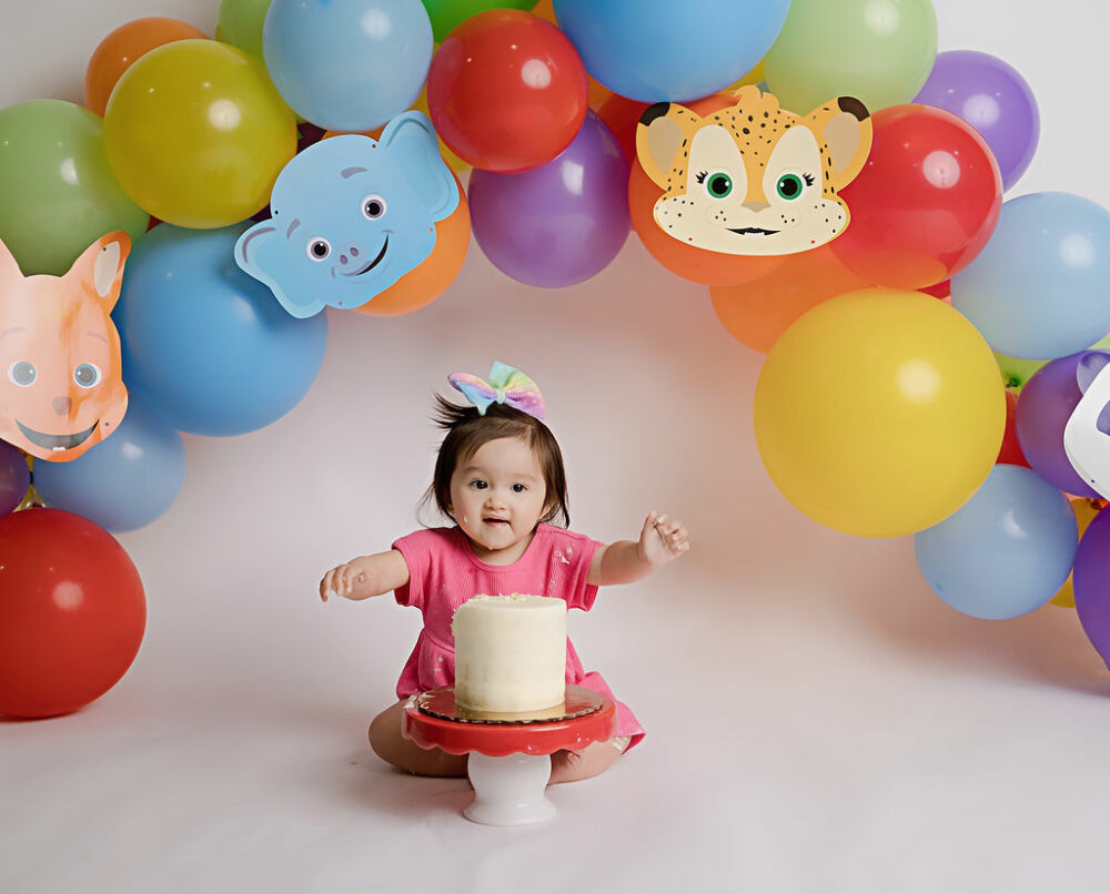 One year old girl sitting on the floor wearing dress and large headband eating cake with her favorite characters as the backdrop for her word party first birthday session taken in Burlington, New Jersey.