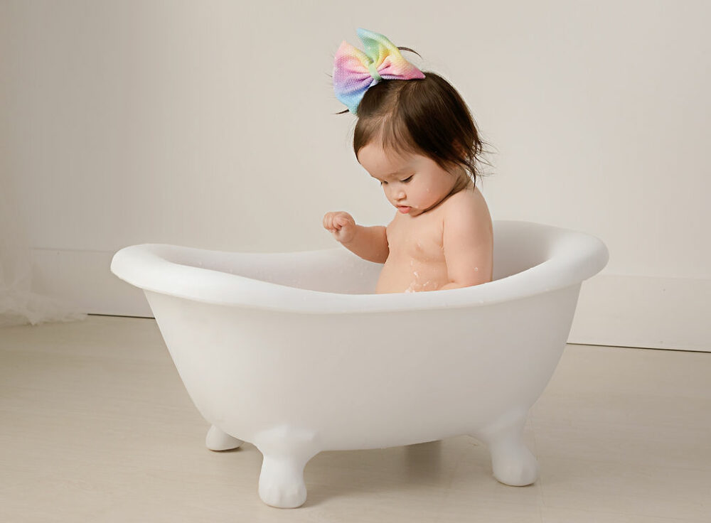 Toddler girl sitting in miniature white tub photography prop wearing large boat taken during her first birthday photo shoot taken in freehold, New Jersey.