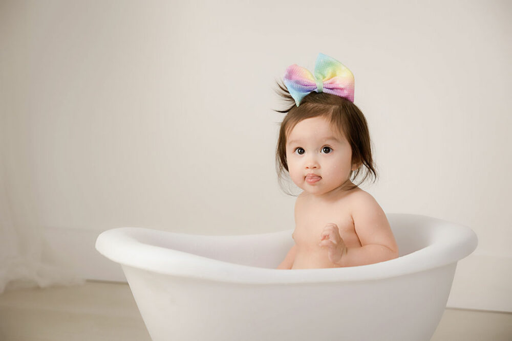 One year old girl sitting in little tub photography prop wearing headband for her professional and studio development photography session taken in Monmouth, New Jersey.
