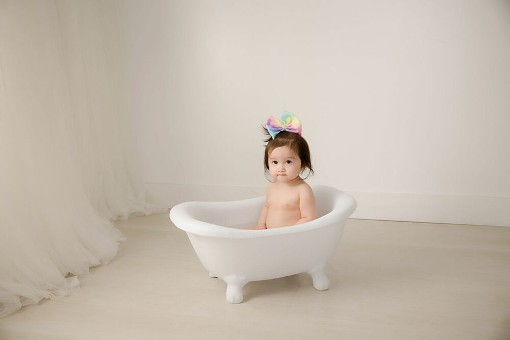 Cute picture of toddler girl in white bathtub photography prop taken during her first birthday photo shoot taken in studio for her word party first birthday session in Camden, NJ.