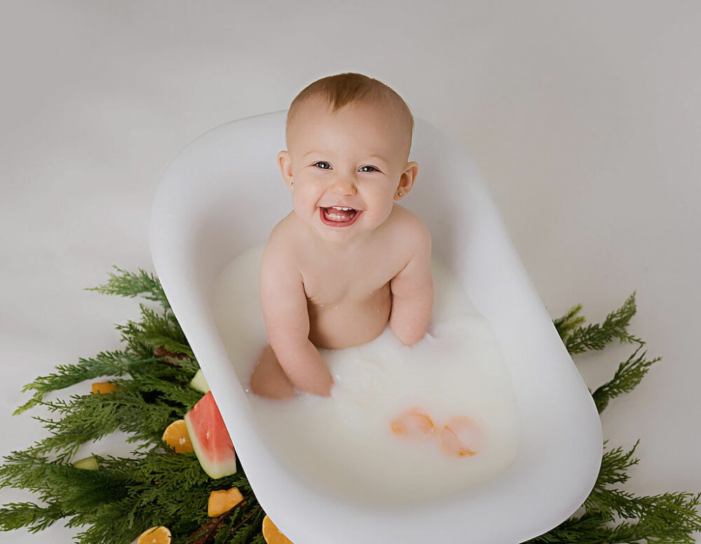 Toddler girl sitting in milk bath with fruit in it, smiling at the camera for her baby mile stone photos taken during her snowflake first birthday session in Wrightstown, New Jersey.