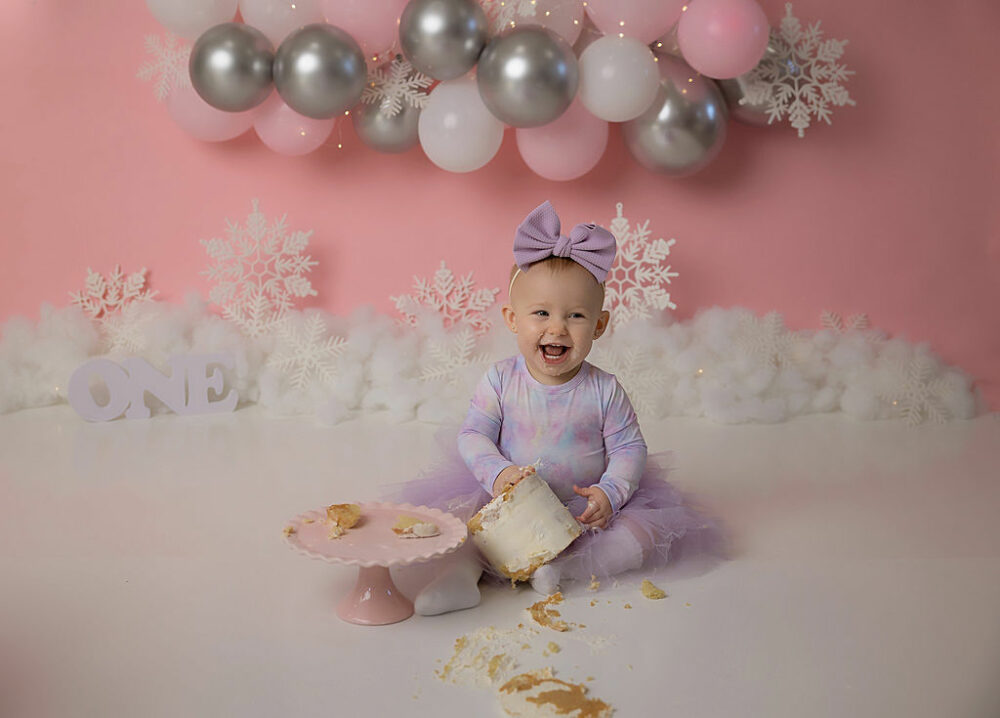 Toddler girl sitting on floor wearing cute outfit and smiling while eating cake for her baby girl first birthday cake smash photo shoot in Hamilton, New Jersey.