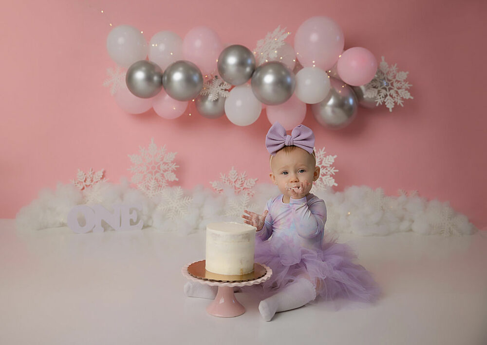 One year old girl in dress, eating her first birthday cake with balloons and fluff and snowflakes as a backdrop for her first birthday session, taken in Cherry Hill, New Jersey.