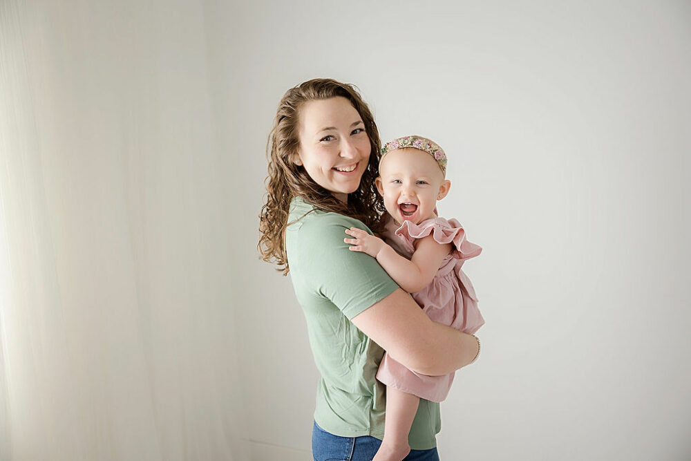 A woman smiling at camera as she is holding her one-year-old daughter, who is also smiling at the camera for a FirstBirthday photoshoot, taken in studio in Mount Holly, New Jersey.
