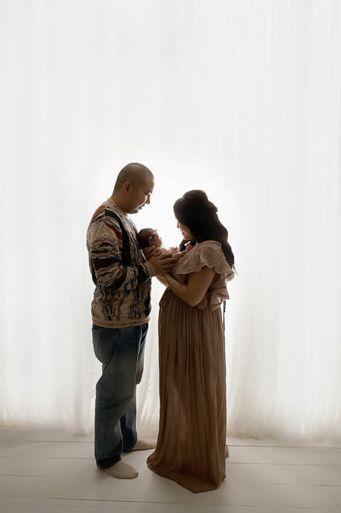 A woman and a man standing facing each other holding their infant between them against a light and bright background for her pink in-home newborn session taken in Southampton, New Jersey.