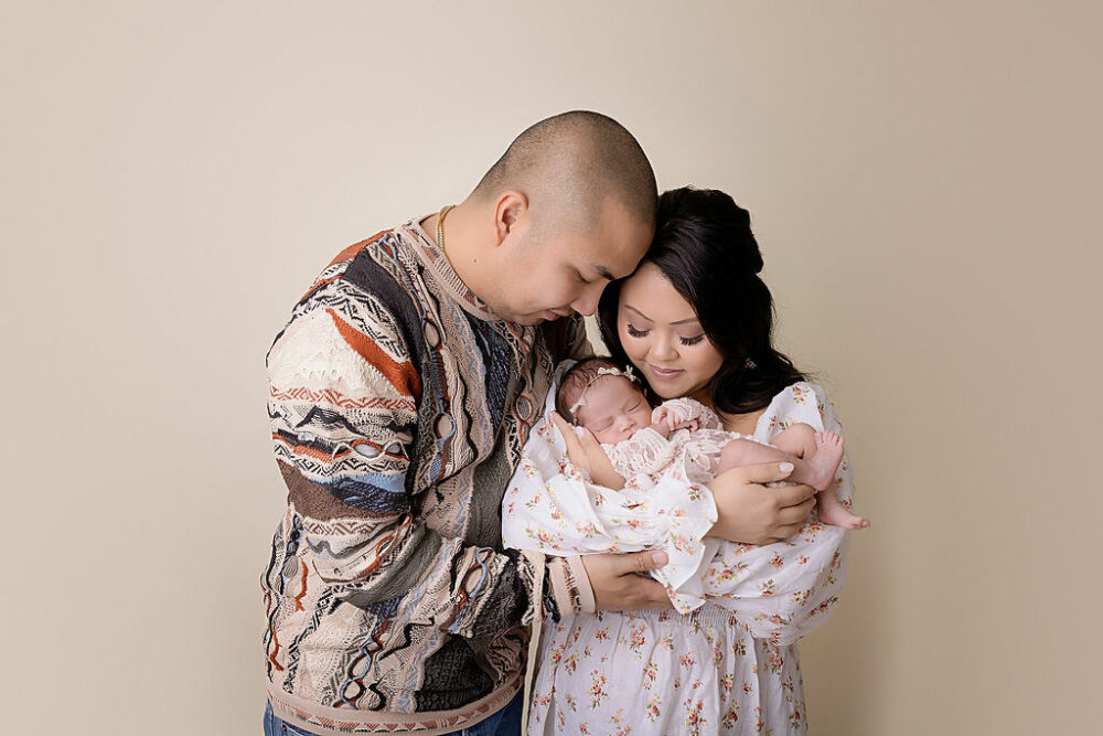 A couple doing couple photography poses as their daughter sleeps in her mothers arms for her pink in-home newborn session in Eastampton, New Jersey.