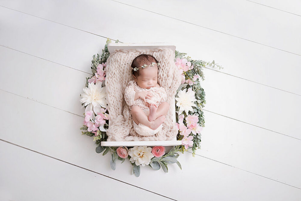A newborn girl sleeping in newborn photography prop adorned with flowers and greenery and blanket for her pink in-home newborn session in Westampton, New Jersey.