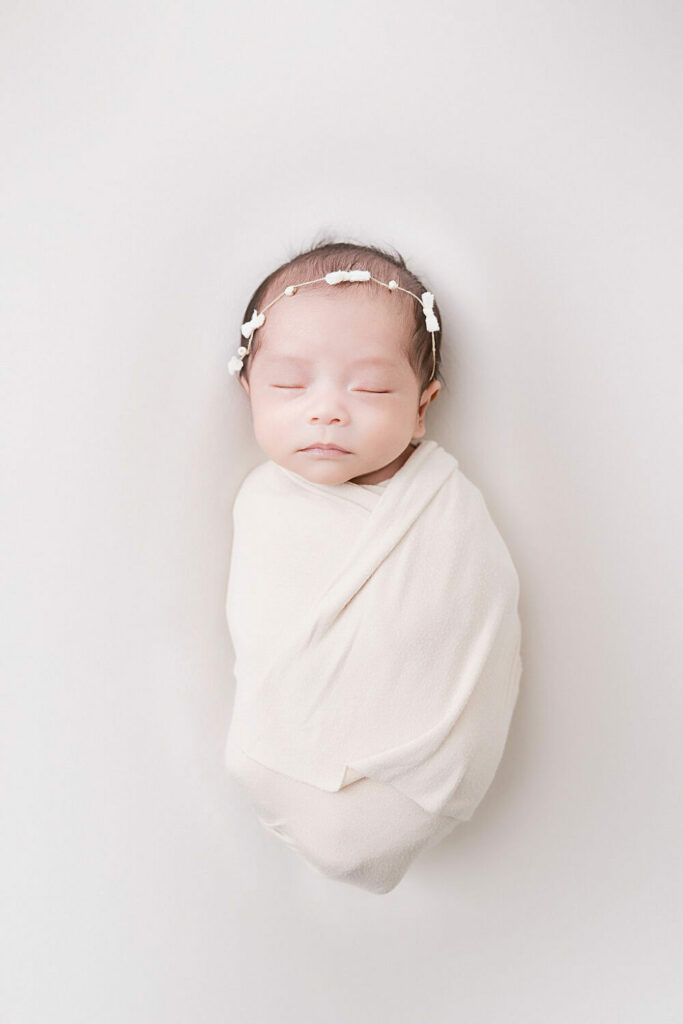 An infant sleeping on her back wrapped in swaddle and wearing dainty headband for her newborn portraits taken in Mount Laurel, New Jersey.