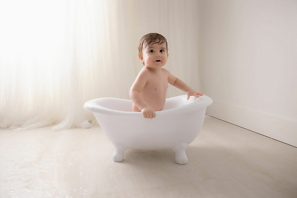 Infant boy sitting in little bathtub photography prop, looking at camera with a light and bright backdrop for his first birthday ideas in Burlington, New Jersey.