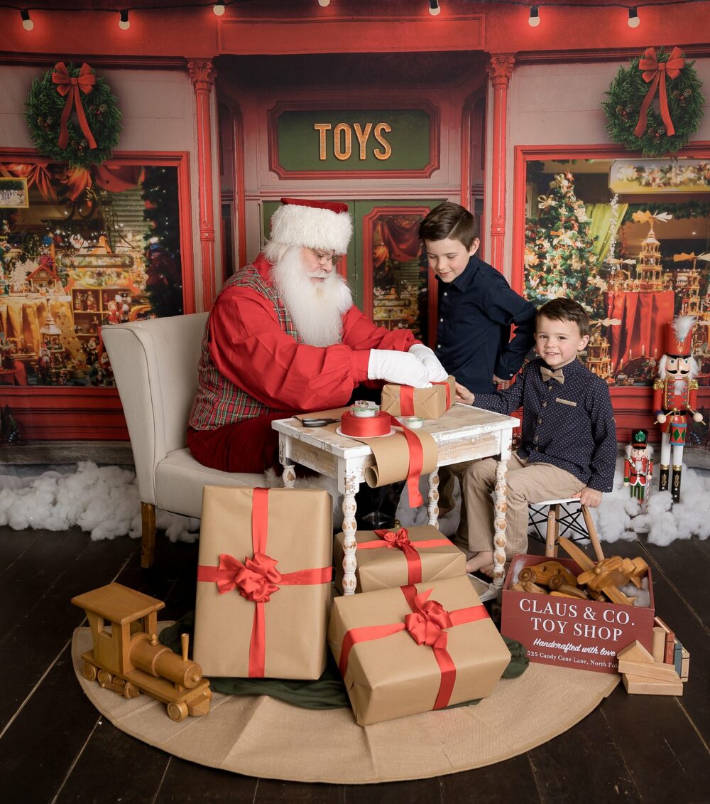 Santa in his toy shop wrapping presents with two boys dressed in blue shirts.