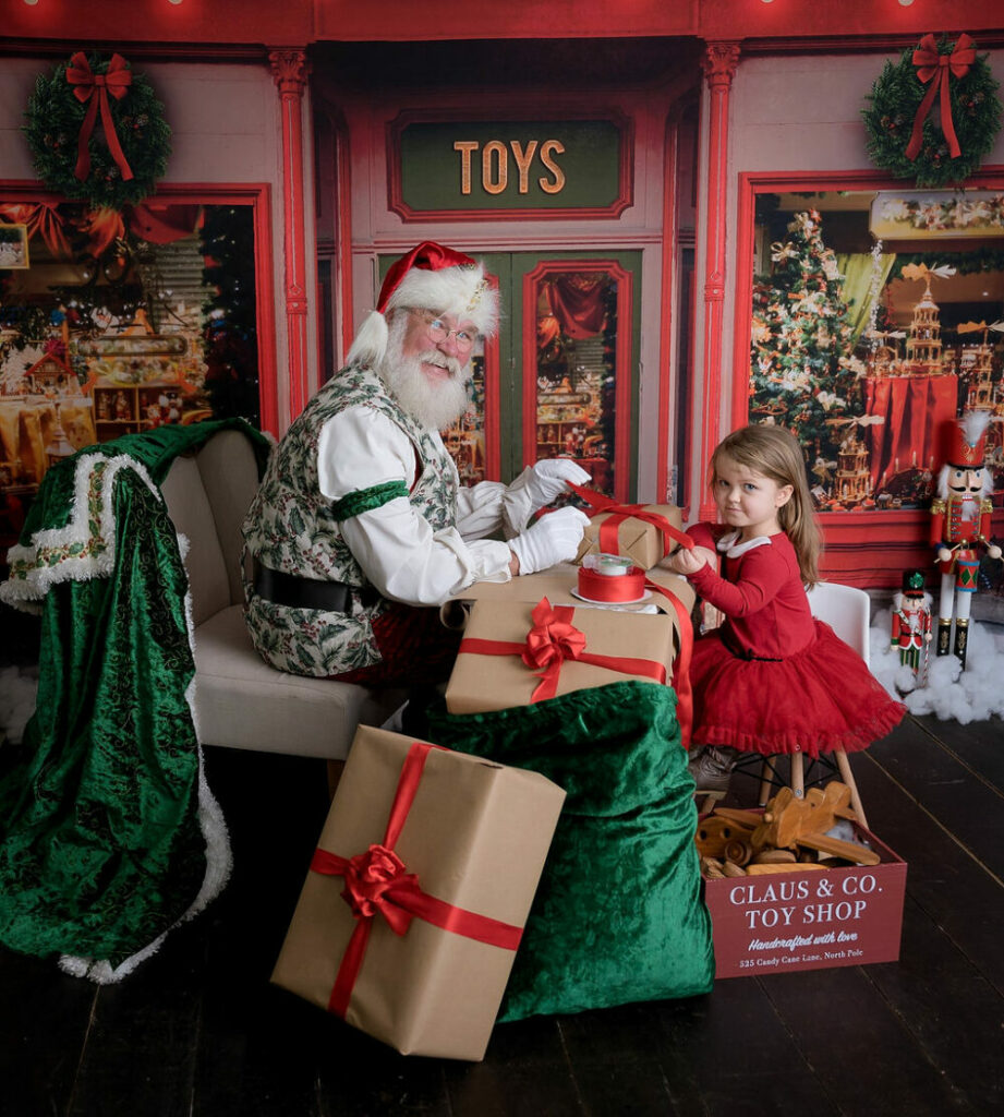 A man dressed up as Santa Claus sitting across from a toddler girl wrapping presents together in a toy shop for their holiday photos sessions in Southampton, New Jersey.