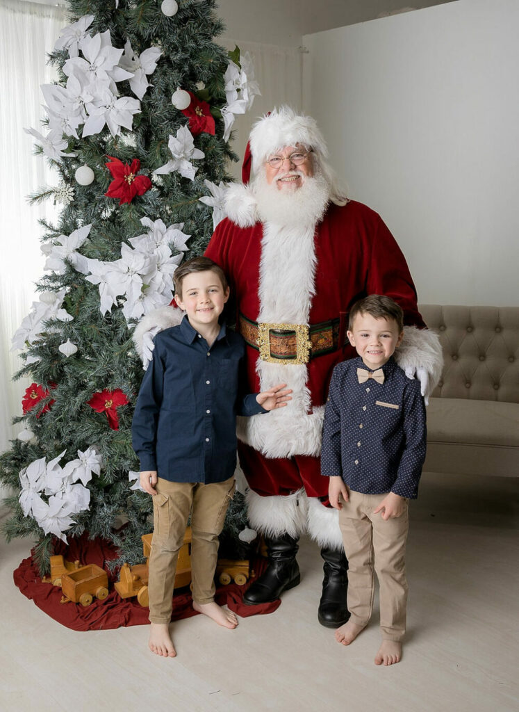 A man dressed up as Santa Claus, wearing boots and hat standing next to two brothers and large Christmas tree for a in studio family photo in Burlington, New Jersey.