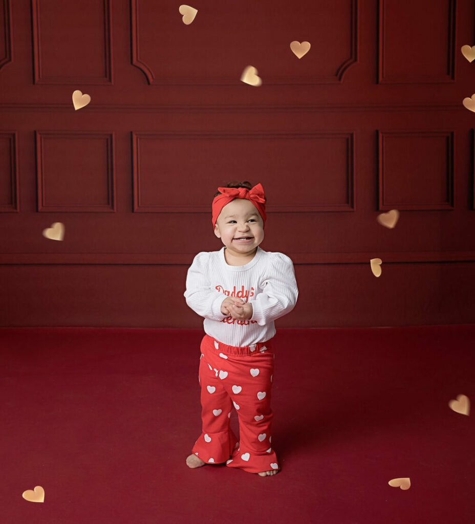 A toddler girl, wearing pants and long sleeve and headband, smiling for her portrait photo taken during Valentine’s Day mini session in Mount Holly, New Jersey.