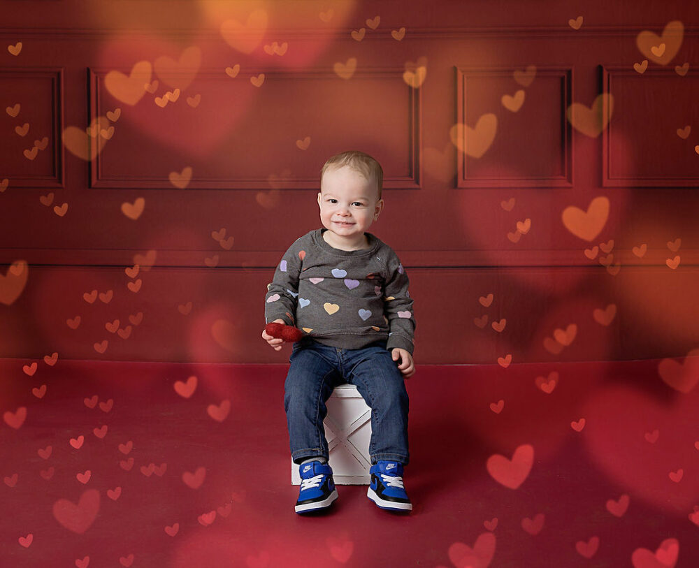 A cute toddler sitting on photography prop holding felt heart and smiling for a Valentines mini photo, shoot in Burlington, New Jersey.