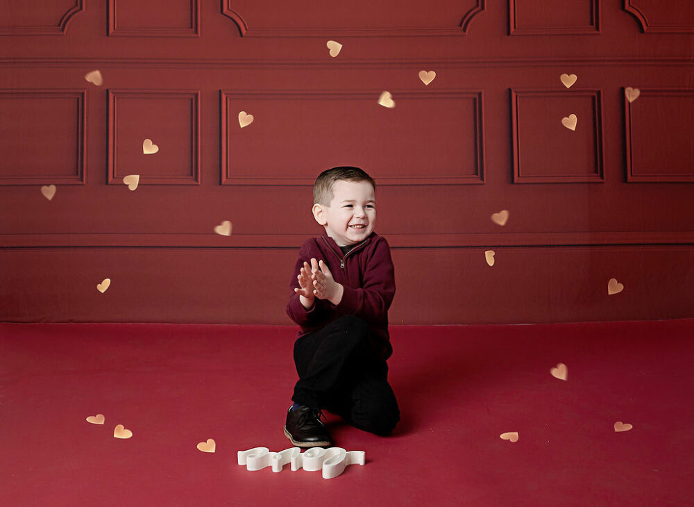 A lifestyle photo of a boy, smiling and sitting on the floor, wearing sweater and pants against a red backdrop for VDay mini sessions in South Jersey.