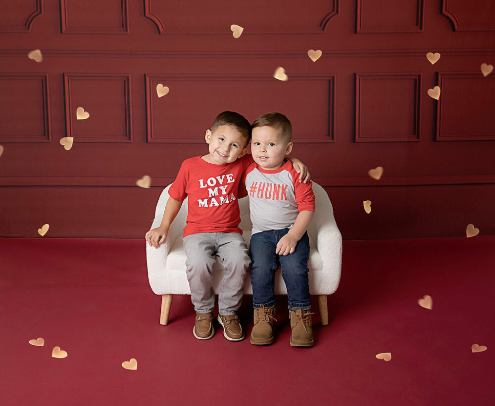 Two toddler brothers sitting on photography prop couch, with their arms wrapped around each other’s shoulders, smiling and wearing tshirts and jeans for a VAlentine’s Day mini session in Eastampton, New Jersey.