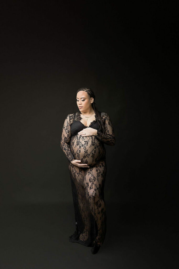A woman doing photography poses wearing lace dress against black backdrop for her maternity portrait, studio photography session in Mount Holly, New Jersey.