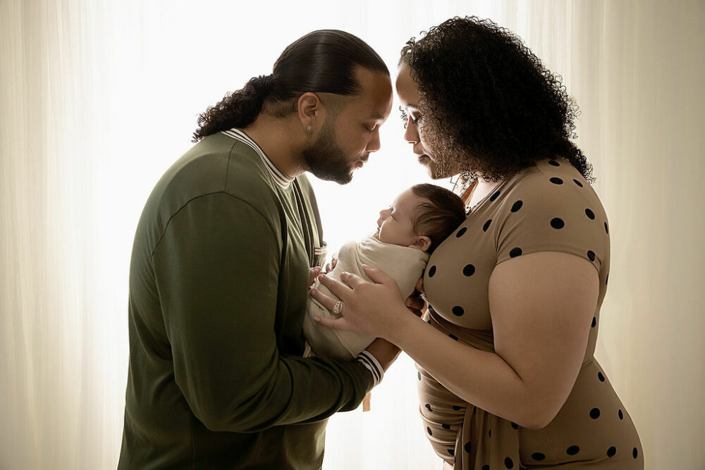 A man and woman facing each other as they hold there anything in between them, almost touching for head against a light and bright background for their couple pregnancy photos taken in Warren, New Jersey.