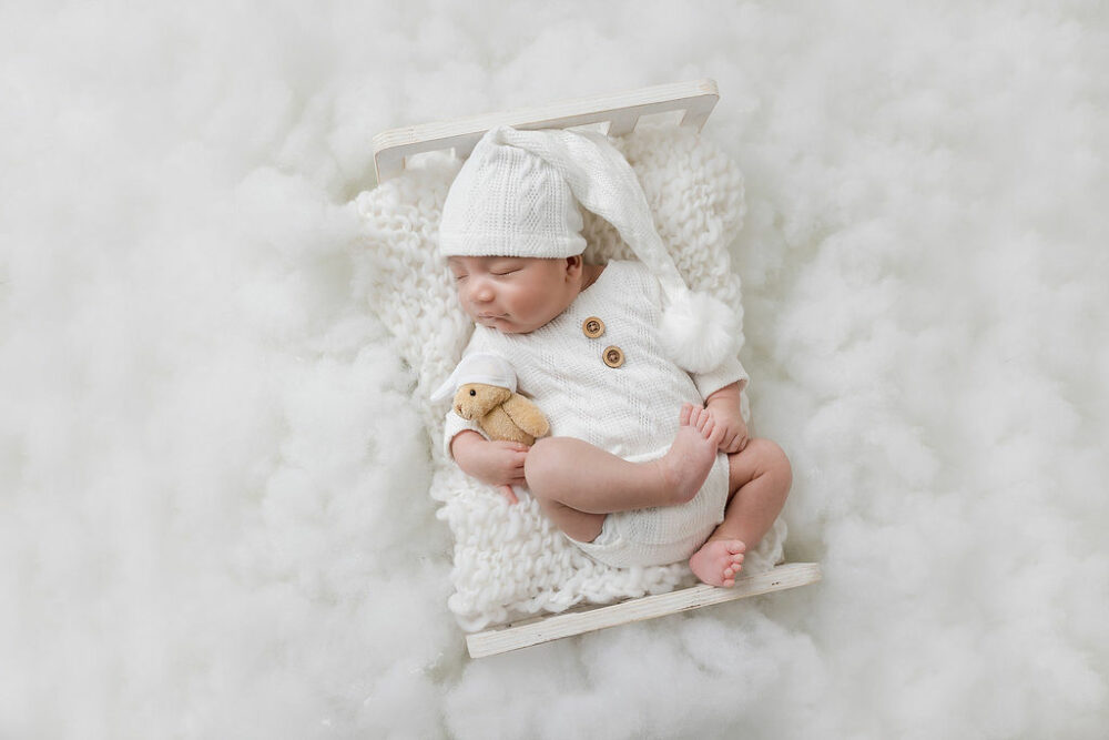 An infant boy, wearing matching outfit and hat, sleeping with teddy bear plush on textured blanket and tiny crib for his in-studio aviation, newborn session in Westampton, New Jersey.