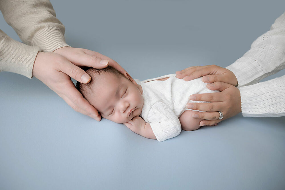 Baby photography of an infant boy sleeping on his tummy, wearing infant outfit, with his parents hands holding him for their in-studio newborn session in Medford, New Jersey.