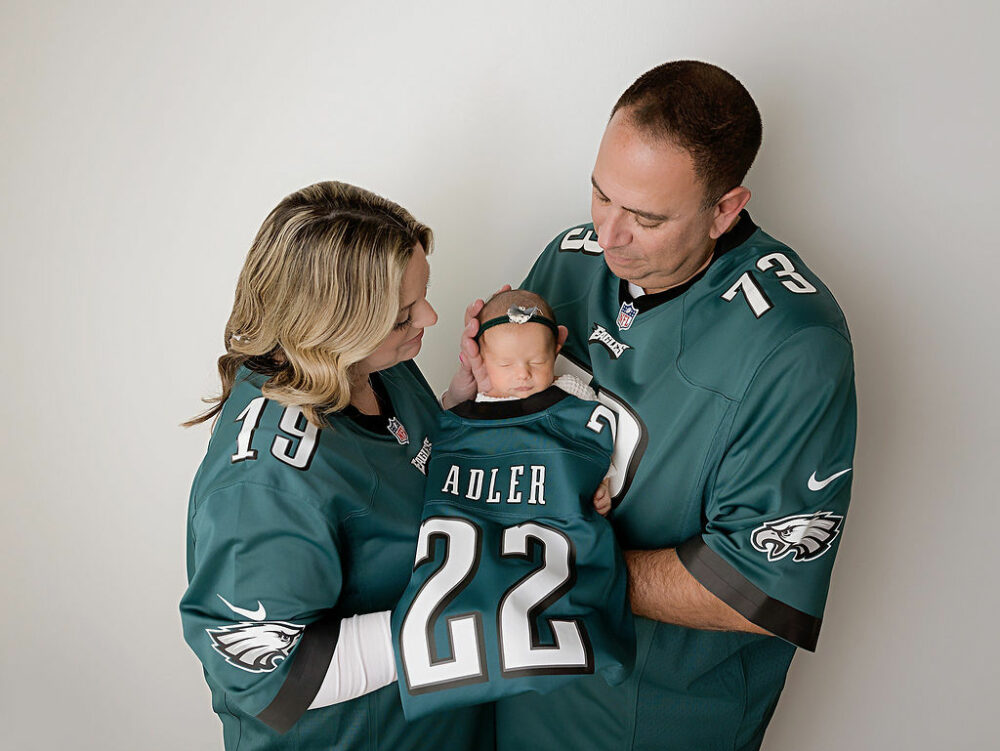 Oh man and a woman holding their newborn daughter in between them well they both wear matching shirts looking at their baby for an Eagles newborn portrait taken in Southampton, New Jersey.